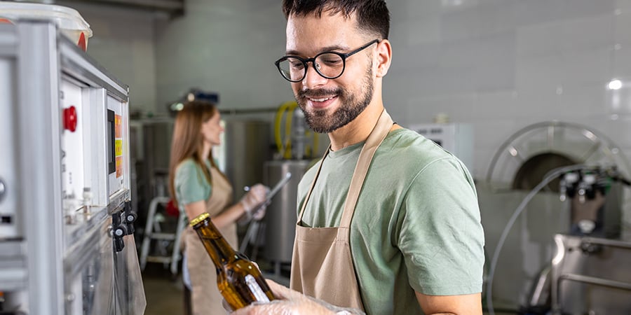 Man with glasses wearing an apron holding a beer bottle coming off a production line